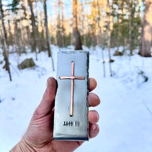 May include: A rectangular metal money clip with a hammered edge finish, featuring a centered copper cross inlay and a stamped tally mark design at the bottom. The surface has a brushed metallic texture with a distinct vertical orientation.