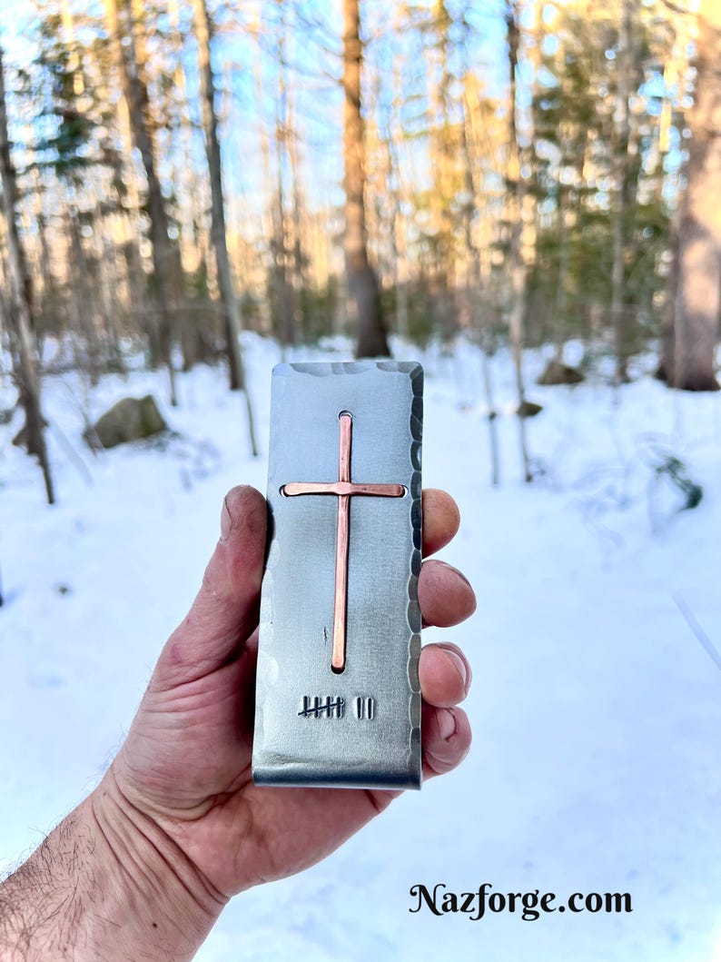 May include: A rectangular metal money clip with a hammered edge finish, featuring a centered copper cross inlay and a stamped tally mark design at the bottom. The surface has a brushed metallic texture with a distinct vertical orientation.