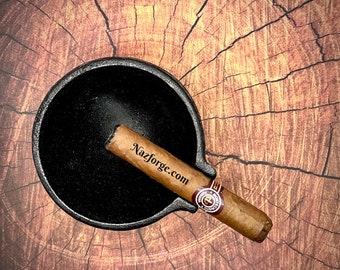 May include: Black cast iron ashtray with a cigar resting on the rim. The cigar is labeled "Nazforge.com".