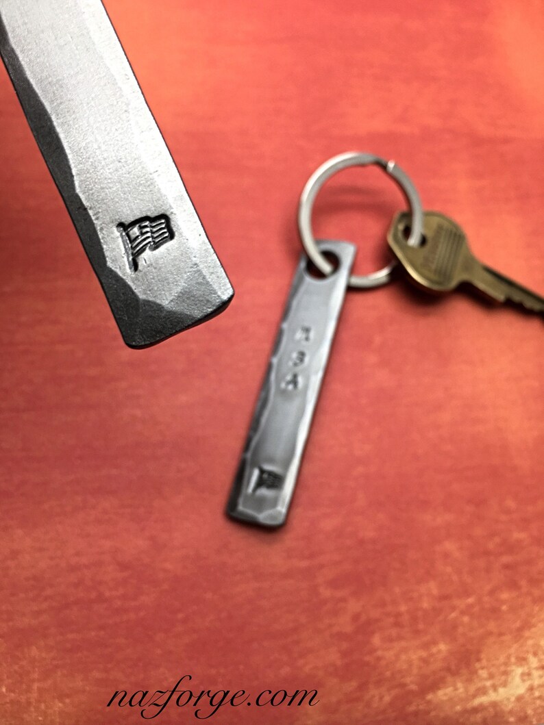 May include: Hand-forged metal keychains with a hammered texture. One keychain features a small American flag emblem. The other keychain has a key ring and a brass key attached. The background is a reddish-orange color.