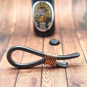 May include: A black metal bottle opener with a copper wire wrap. The opener has a loop on one end and a bottle opener on the other end.