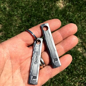 May include: Two rectangular, textured metal keychains. One keychain has the letters "USA" and a flag symbol stamped on it. The other keychain has a flag symbol stamped on it. Both keychains have a hole at the top for a keyring.