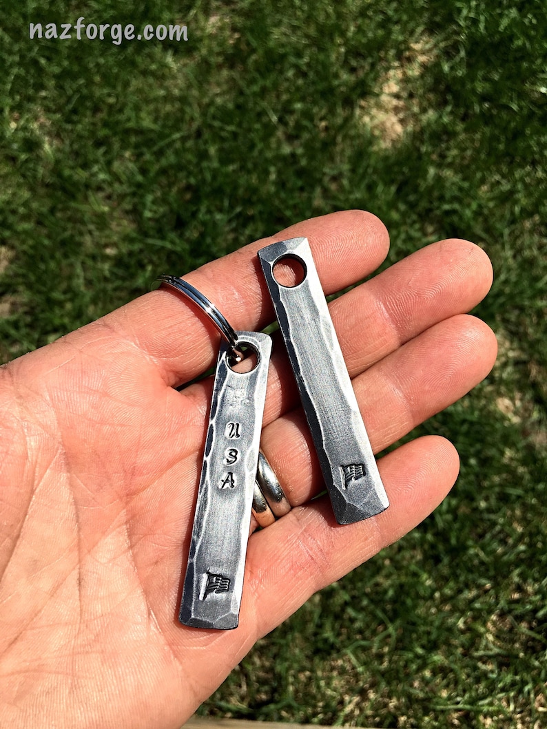 May include: Two rectangular, textured metal keychains. One keychain has the letters "USA" and a flag symbol stamped on it. The other keychain has a flag symbol stamped on it. Both keychains have a hole at the top for a keyring.