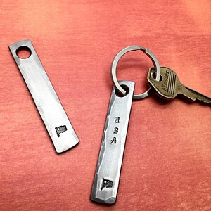 May include: Two metal keychains with a hammered finish. One keychain has a small American flag and the other has "USA" stamped on it, along with a small flag. A key is attached to the keychain. The website nazforge.com is in the top of the image.