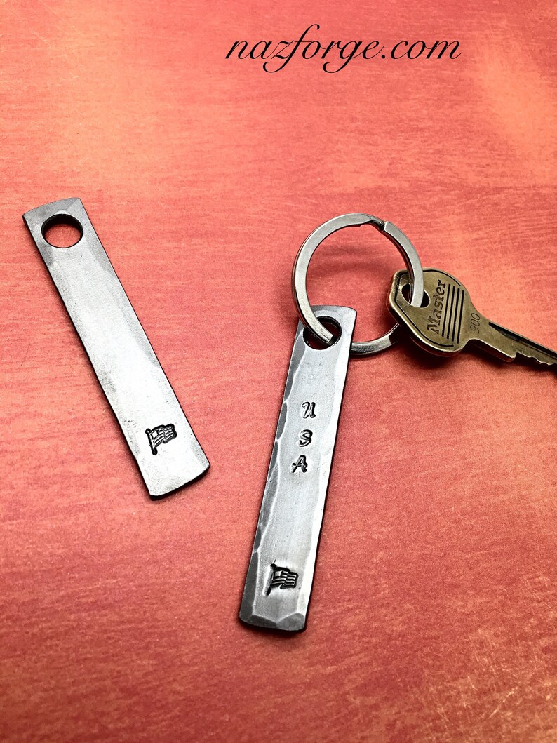 May include: Two metal keychains with a hammered finish. One keychain has a small American flag and the other has "USA" stamped on it, along with a small flag. A key is attached to the keychain. The website nazforge.com is in the top of the image.