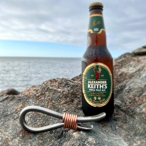 May include: A silver bottle opener with a copper wire wrap, sitting on a rock next to a bottle of Alexander Keith's India Pale Ale.