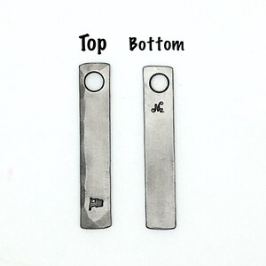 May include: Two rectangular metal pendants with a hammered texture. One pendant has a small flag design, the other has an initial. Both have a hole at the top. The words "Top" and "Bottom" are above the pendants. The website nazforge.com is at the bottom.