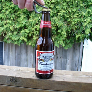 May include: A hand using a metal bottle opener to open a bottle of Budweiser beer. The bottle is on a wooden surface.