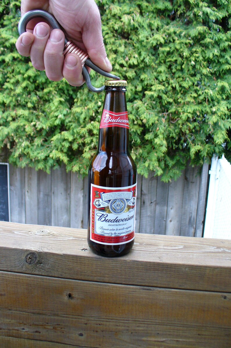 May include: A hand using a metal bottle opener to open a bottle of Budweiser beer. The bottle is on a wooden surface.