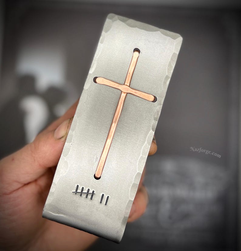 May include: A rectangular silver-toned metal money clip with a hammered texture along the edges. It features an inlaid copper cross in the center and a stamped tally mark design consisting of five horizontal lines and two vertical lines at the bottom.