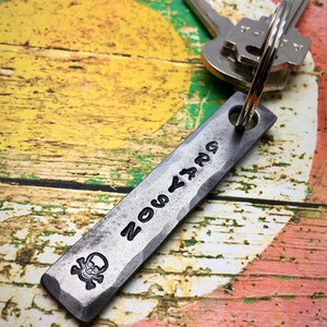 May include: A silver metal keychain with a skull and crossbones design and the word "ZOMBIE" engraved on it. The keychain is attached to a set of keys.