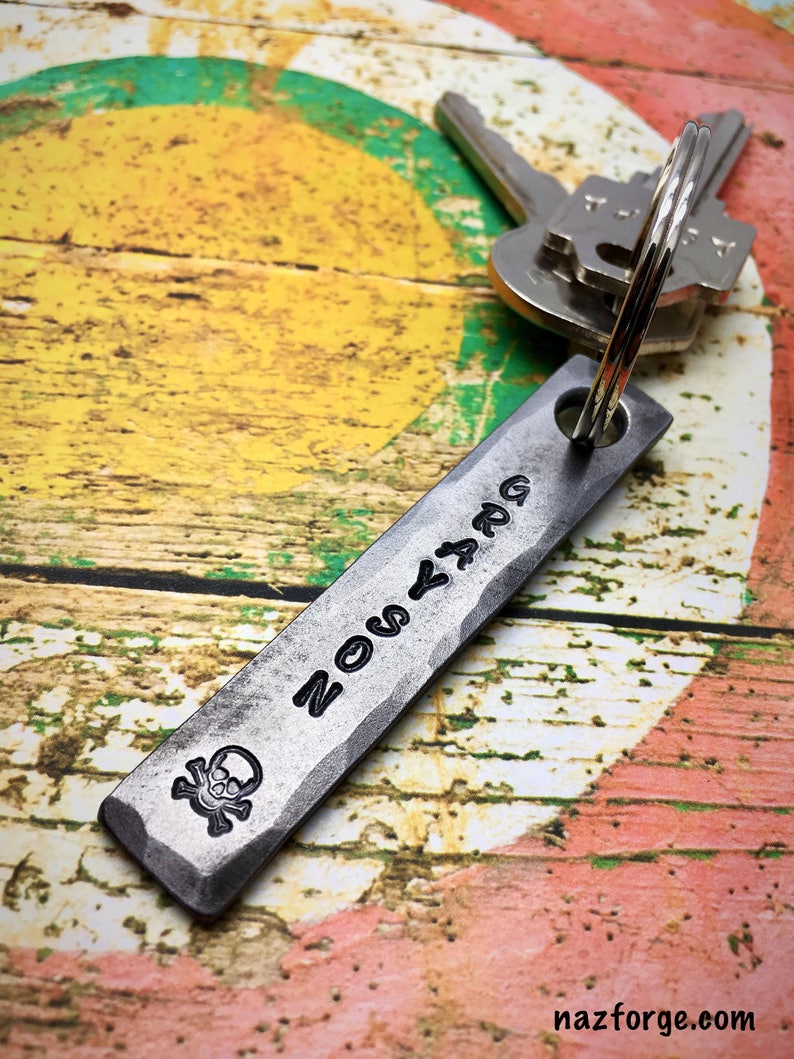 May include: A silver metal keychain with a skull and crossbones design and the word "ZOMBIE" engraved on it. The keychain is attached to a set of keys.