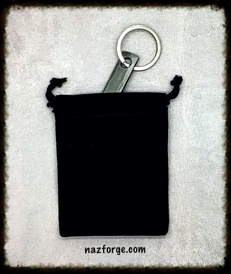 May include: A silver key chain with a small, flat, rectangular blade attached to a silver ring. The key chain is in a black velvet pouch with a drawstring.