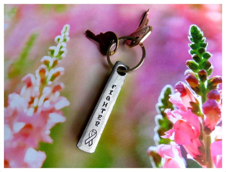May include: Silver key chain with a charm that says "Fighter" and a pink ribbon. The keychain is on a pink and green floral background.
