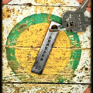 May include: A silver metal keychain with the name "GRAYSON" engraved on it. The keychain has a skull and crossbones design and is attached to a key with a silver metal ring. The keychain is on a wooden surface with a green, yellow, and red painted circle.