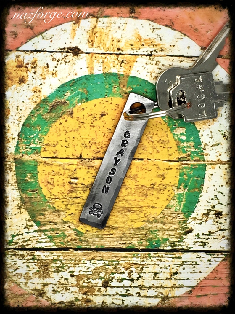 May include: A silver metal keychain with the name "GRAYSON" engraved on it. The keychain has a skull and crossbones design and is attached to a key with a silver metal ring. The keychain is on a wooden surface with a green, yellow, and red painted circle.