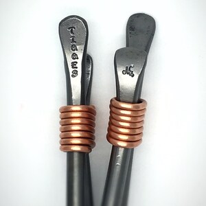 May include: Two silver metal hair clips with copper wire wrapped around the middle. The top of the clips are engraved with the words "TIGGES" and "Naz".