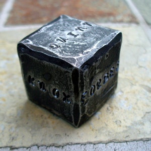 May include: A metal cube with the number "1" and the letters "K.M.G." engraved on its sides. The cube has a weathered, aged appearance.