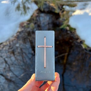 May include: A rectangular, hammered-finish steel money clip featuring a centered, vertical copper cross inlay. The metal surface displays a textured, rustic appearance. The text Nazforge.com is visible in the upper left corner of the image.