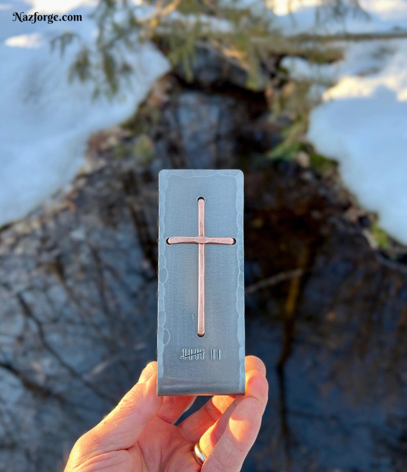 May include: A rectangular, hammered-finish steel money clip featuring a centered, vertical copper cross inlay. The metal surface displays a textured, rustic appearance. The text Nazforge.com is visible in the upper left corner of the image.
