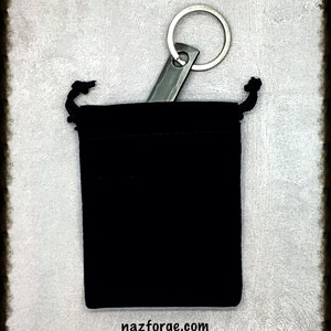 May include: A black velvet drawstring pouch with a silver metal keychain. The keychain has a rectangular tag attached to a circular ring. The website nazforge.com is printed at the bottom of the image.