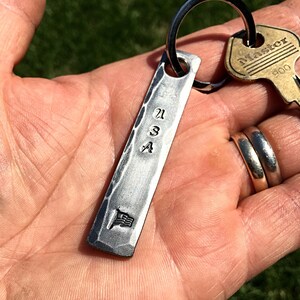 May include: A silver-colored rectangular keychain with the letters "USA" stamped on it, along with a small American flag emblem. The keychain is attached to a key ring with a "Master" key. The website nazforge.com is visible in the background.