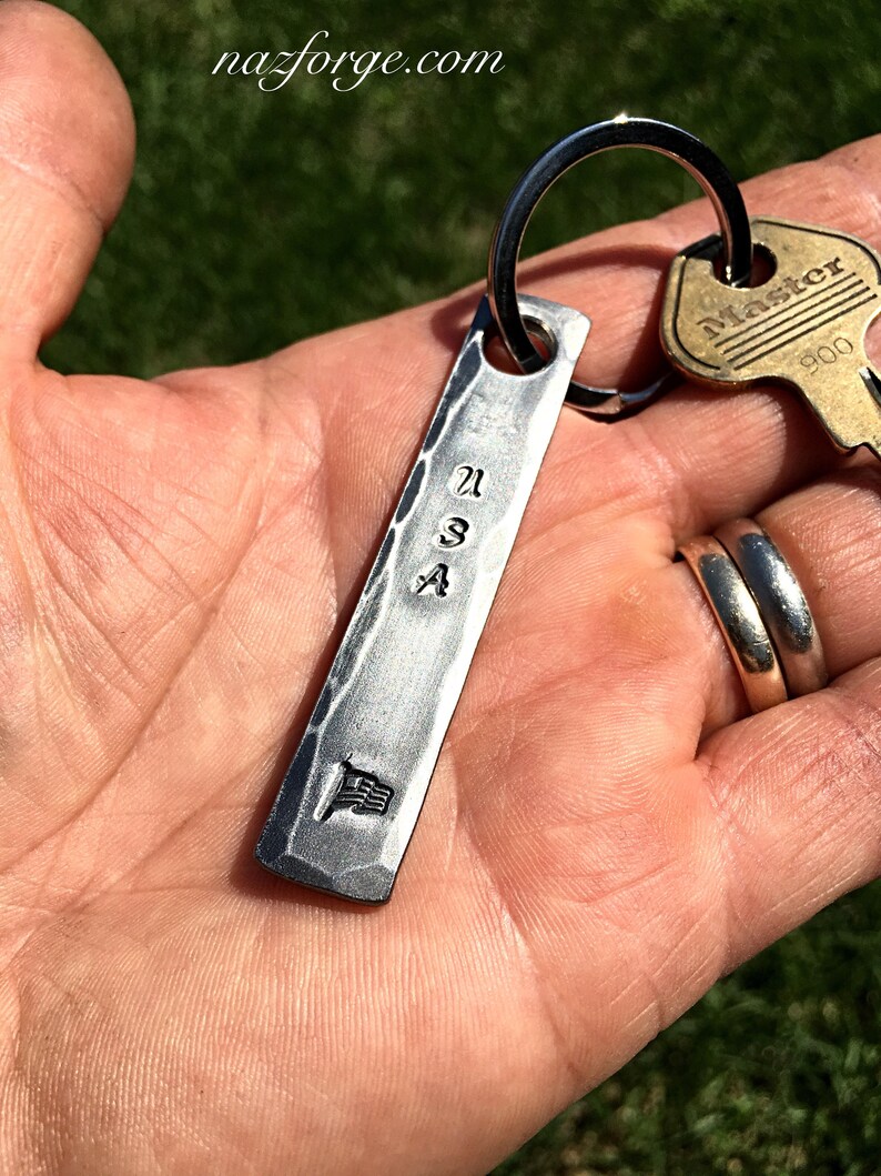 May include: A silver-colored rectangular keychain with the letters "USA" stamped on it, along with a small American flag emblem. The keychain is attached to a key ring with a "Master" key. The website nazforge.com is visible in the background.