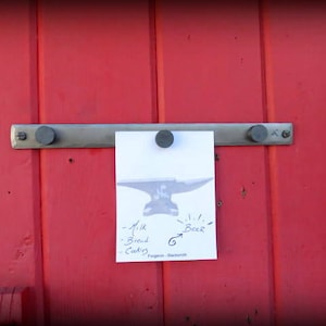 May include: A white card with a black and white illustration of an anvil and the text "Milk, Bread, Cookies, Beer" is attached to a metal bar with two black knobs on a red wooden door.