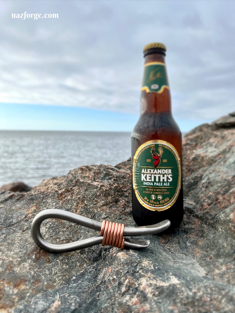 May include: A silver bottle opener with a copper wire wrap, sitting on a rock next to a bottle of Alexander Keith's India Pale Ale.