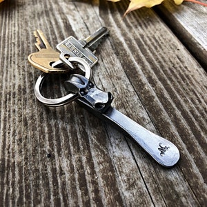 Puede incluir: Un llavero de plata con un pequeño charm de metal con la cabeza de un dragón detallada. El charm tiene una pequeña letra "M" grabada en la parte inferior. El llavero está sobre una superficie de madera con una hoja amarilla en el fondo.