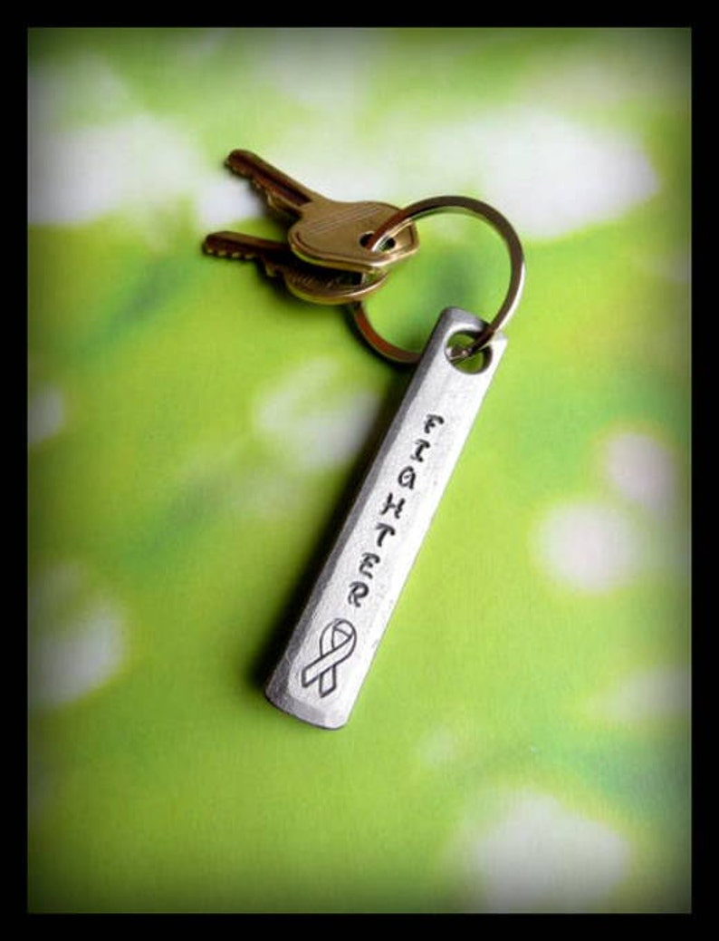 May include: Silver keychain with a silver tag that reads "Fighter" with a ribbon design. The keychain is attached to two gold keys.