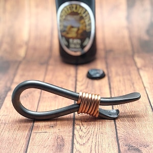 May include: A black metal bottle opener with a copper wire wrap. The opener has a loop on one end and a bottle opener on the other end.