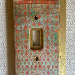 Könnte beinhalten: Dekorative Lichtschalterabdeckung mit goldfarbenem Rahmen. Die Mitte zeigt ein gemustertes Design in Türkis, Rot und Gold. Die Abdeckung hat eine einzelne Schalteröffnung und zwei Schraubenlöcher.