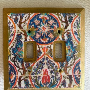 May include: A square double light switch wall plate featuring a gold-painted border and a central panel decorated with a colorful floral pattern in shades of blue, orange, and white. The plate includes two rectangular cutouts for switches.
