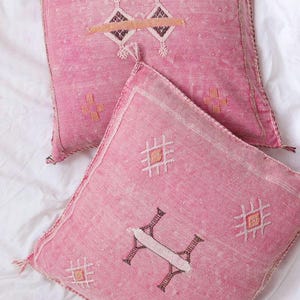 May include: Two pink square throw pillows with geometric designs. The pillows have a textured appearance and feature embroidered patterns in white, brown, and orange. Tassels are visible on the corners.