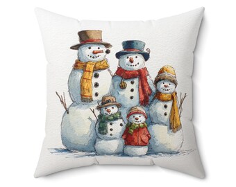 Op de afbeelding: Een wit vierkant kussen met een aquarel illustratie van een familie sneeuwmannen. De sneeuwmannen dragen hoeden, sjaals en hebben takken als armen. Het kleurenpalet omvat wit, oranje, rood, groen en bruin. Het kussen heeft een wintervakantiethema.