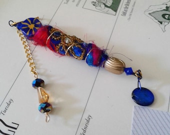 May include: A decorative hanging ornament featuring a central cylindrical component wrapped in blue and magenta fibers, accented with gold wire, blue beads, and a metallic gold bead. A gold chain with iridescent beads and a blue disc charm hangs from it.