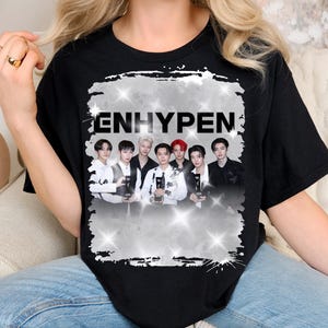 Peut inclure: Un t-shirt noir à manches courtes avec une impression graphique centrale du groupe de K-pop ENHYPEN. Le motif comprend le texte ENHYPEN en lettres noires grasses sur une photo de groupe, le tout sur un fond blanc usé avec des effets d'étoiles.