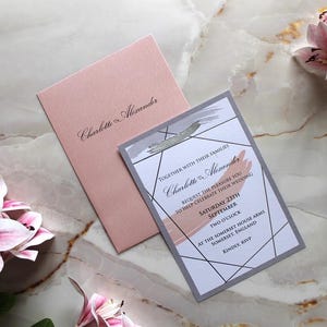 Può includere: Un set di partecipazioni di nozze con una busta rosa e un biglietto d'invito abbinato. Il biglietto presenta disegni geometrici, testo e accenti di pennellate. Il testo recita "Charlotte & Alexander" e i dettagli del matrimonio. Fiori rosa e bianchi sullo sfondo.