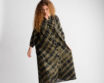 May include: A long-sleeved, button-down dress with a tie-dye pattern in shades of olive green and black. The dress has a mandarin collar and is worn with brown shoes. The dress is knee-length.