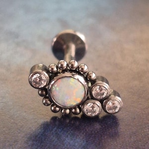 Peut inclure: Un bijou de piercing labret argenté avec une pierre centrale opalescente, entourée de petits cristaux clairs et de minuscules perles argentées. Le bijou est posé sur un fond flou et multicolore.