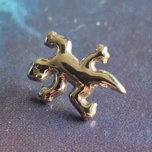 May include: A small, shiny silver lizard pin. The lizard is stylized with a smooth, reflective surface, and is posed with its legs spread. The pin is set against a blue and purple background.