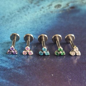 May include: Five silver ear studs with a flat top, each featuring a triangular cluster of gemstones. The gemstones are purple, pink, blue, green, and clear. The studs are presented against a blue and teal background.