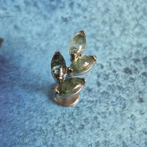 May include: A silver ring with four oval green gemstones. The gemstones have visible inclusions and are set in a prong setting. The ring is displayed against a blue background. The gemstones appear to be approximately 1 cm in length.