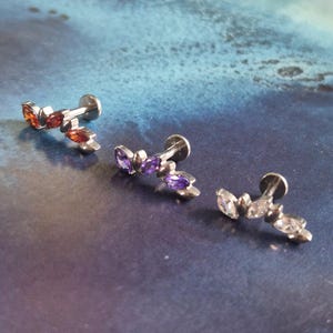 May include: Three silver-toned ear studs, each featuring a row of marquise-cut gemstones. One stud has orange and red stones, another has purple stones, and the last has clear stones. The studs are displayed on a blue and teal background.