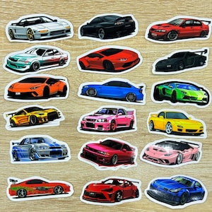 Op de afbeelding: Een verzameling gestanste autostickers met verschillende sportwagenontwerpen in verschillende kleuren. De stickers bevatten auto's in tinten rood, zwart, blauw, groen, geel en roze. De stickers liggen op een lichtbruin oppervlak.