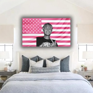 Könnte beinhalten: Ein gerahmtes Wandbild mit einem Schwarz-Weiß-Porträt einer lächelnden Person, vor einem rosa amerikanischen Flaggen-Design. Das Kunstwerk wird über einem Bett mit grauer Bettwäsche und dekorativen Kissen ausgestellt.