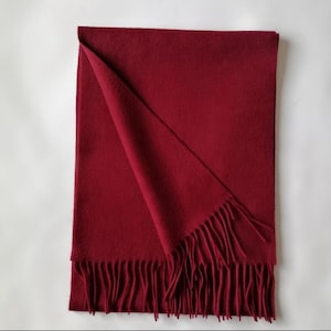 May include: A folded, deep red scarf with fringed edges. The scarf is made of a soft material and is displayed on a white background. The scarf is rectangular in shape and has a simple, classic design.