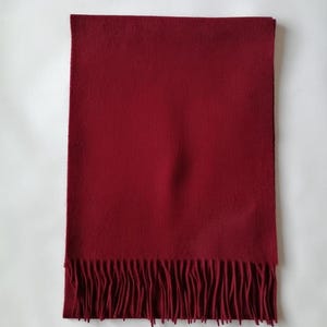 May include: A rectangular, solid burgundy scarf with a fringed edge. The scarf is made of a soft material and is displayed on a white background. The scarf is a winter accessory.