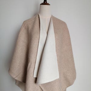 May include: A beige cape-style wrap with a contrasting off-white interior, displayed on a mannequin. The wrap has a draped design with a wide collar and a soft, textured appearance. The neutral color palette suggests versatility and a focus on comfort.